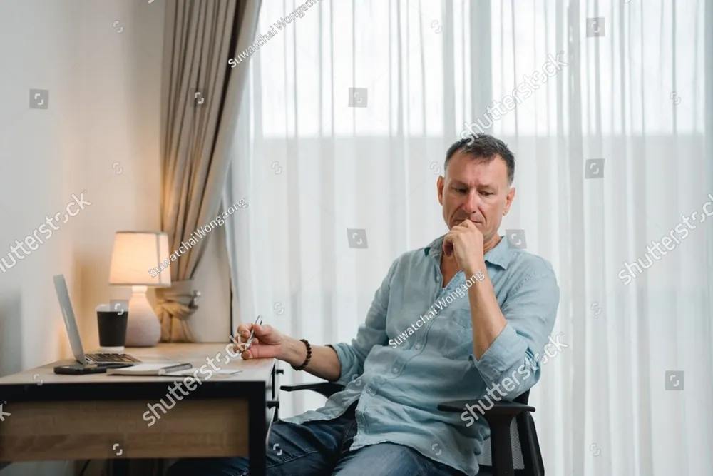 A man at his desk by a bright window, his hand at his chin