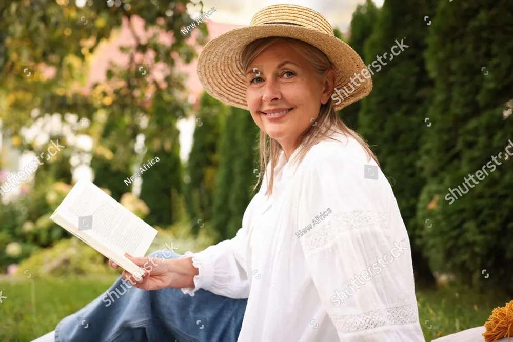 A smiling woman in a straw hat reading a book in a garden