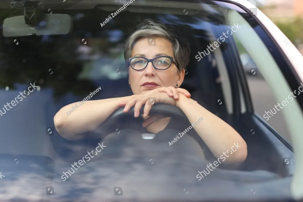 A woman in glasses resting her arms on the open window of a car