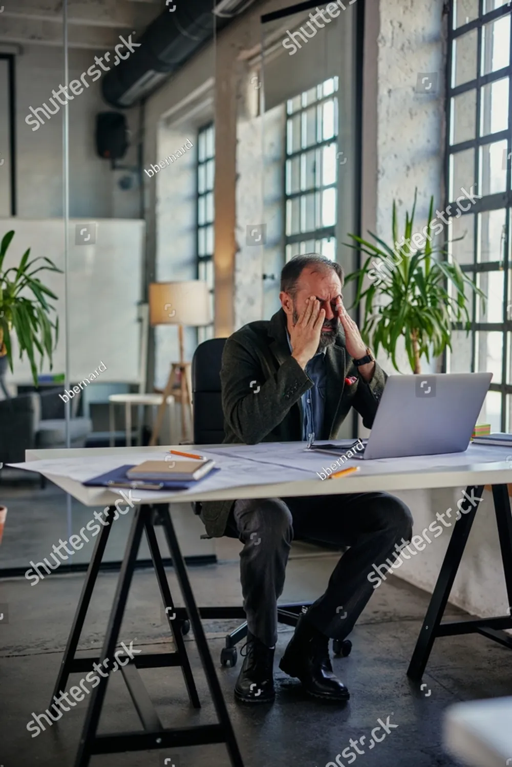 A man at his laptop in an open office, his head in his hand