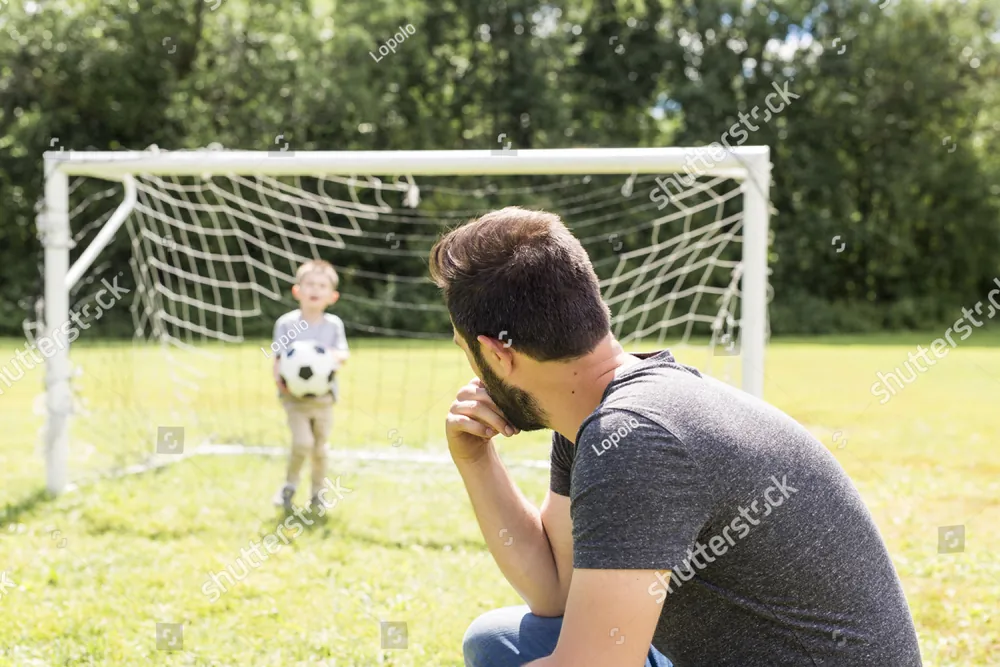 A father kneeling on a soccer field while his young son holds the ball by the goal