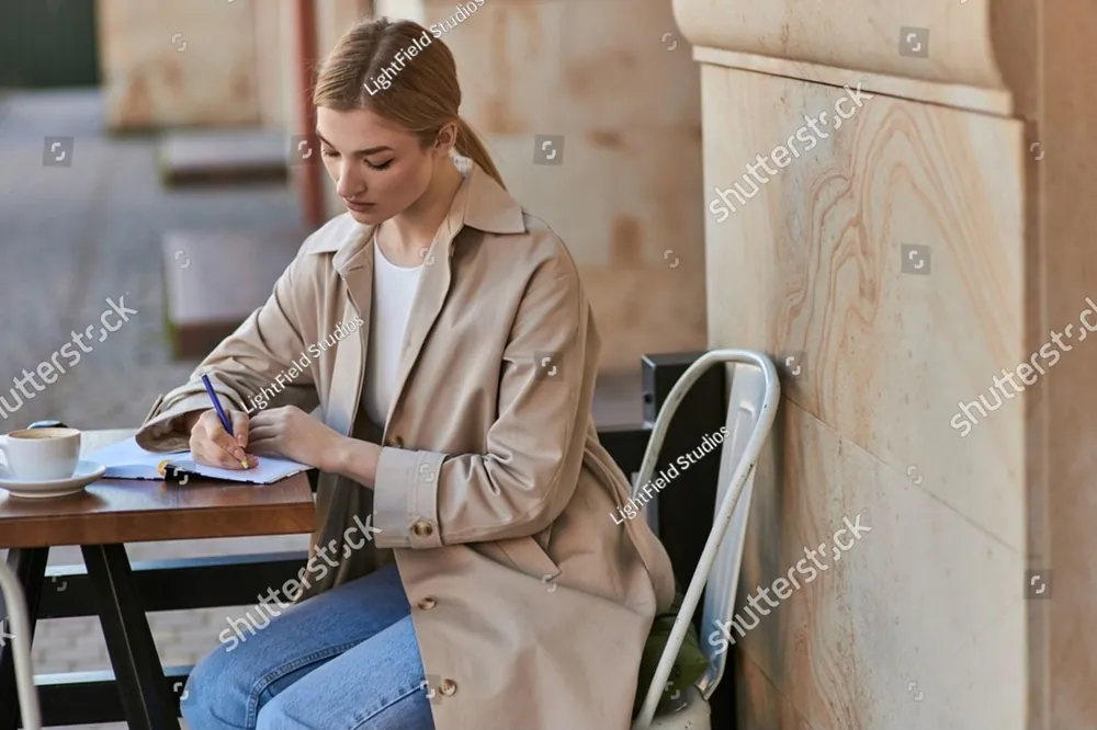 A woman in a trench coat writing in a notebook at a cafe table